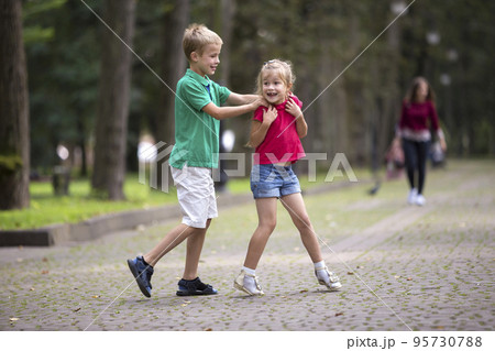 Two cute young laughing children, girl and boy, brother and sister having fun on blurred bright sunny summer park alley green trees bokeh background. Games and recreation, happy childhood concept. 95730788