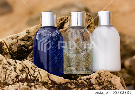 Plastic bottles with natural cosmetic in travel containers - blue shower gel, shampoo with bubbles inside, body lotion in miniature containers against a brown stone rock background. Face body care. 95732339