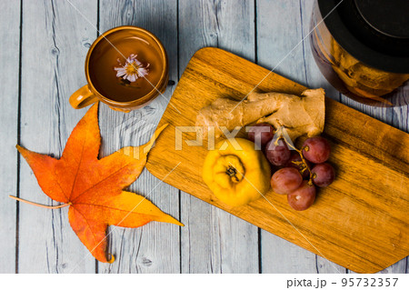 Autumnal still life - bunch of grapes, ripe quince, ginger root lie on wooden cutting board top view. Orange maple leaf on table. Yellow cup with hot aromatic tea drink decorated with flower. 95732357