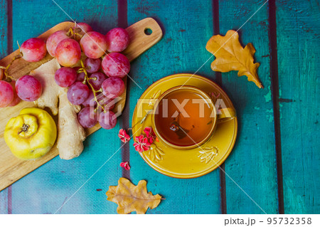 Beautiful fall still life with yellow cup on saucer. Bunch of red grapes, ripe agave, ginger root lie on cutting board. Fallen oak, maple leaves. Wooden green emerald background top view flatly. Beautiful fall still life with yellow cup on saucer. Bunch of red grapes, ripe agave, ginger root lie on cutting board. Fallen oak, maple leaves. Wooden green emerald background top view flatly. 95732358
