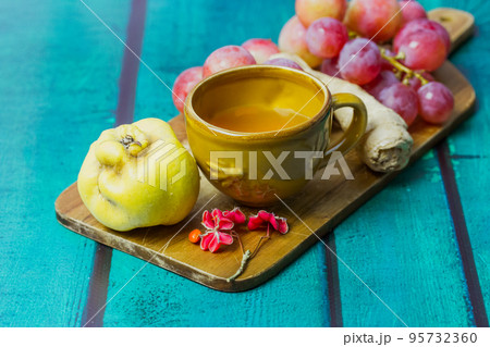 Beautiful fall still life with yellow cup on saucer. Bunch of red grapes, ripe agave, ginger root lie on cutting board. Fallen oak, maple leaves. Wooden green emerald background with space for text. Beautiful fall still life with yellow cup on saucer. Bunch of red grapes, ripe agave, ginger root lie on cutting board. Fallen oak, maple leaves. Wooden green emerald background with space for text. 95732360