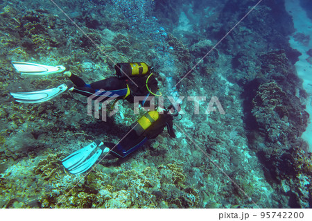 Underwater photo - unknown beginner diver swimming above ocean floor with corals, instructor near holding his oxygen tank, view from above 95742200