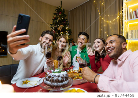 Diverse friends celebrating Christmas and New Year in the evening, sitting at the table, drinking wine and eating, taking a selfie together on the phone near the Christmas tree. Diverse friends celebrating Christmas and New Year in the evening, sitting at the table, drinking wine and eating, taking a selfie together on the phone near the Christmas tree. 95744459