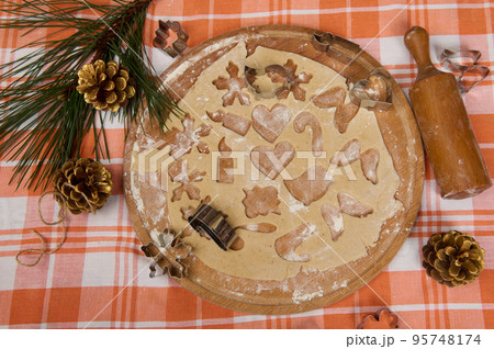 Top view of rolled out gingerbread dough on a wooden board sprinkle with flour, on the table with red plaid tablecloth and pine branch, cones and rolling pin. Making festive pastries. Merry Christmas Top view of rolled out gingerbread dough on a wooden board sprinkle with flour, on the table with red plaid tablecloth and pine branch, cones and rolling pin. Making festive pastries. Merry Christmas 95748174