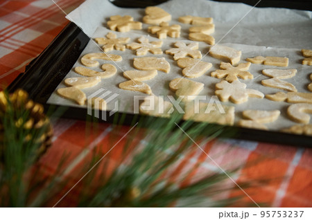 View through blurred fir-tree needles and golden pine cones, to a baking sheet with carved molds of gingerbread dough before baking in the oven. Christmas background. Still life. Festive pastries 95753237