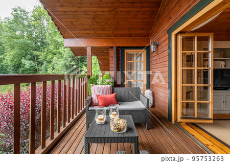Cozy terrace with table and sofa at wooden cottage house surrounded by greenery and trees in countryside on cloudy summer day 95753263