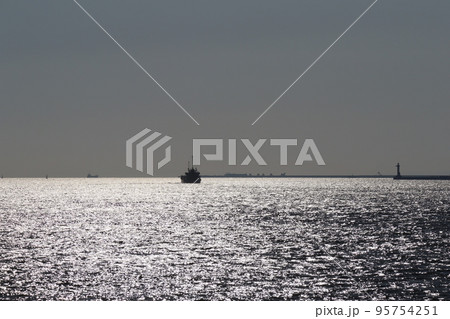 青空と水平線とキラキラ光る海面の海と船と灯台と突堤などがシルエットで見える風景 95754251