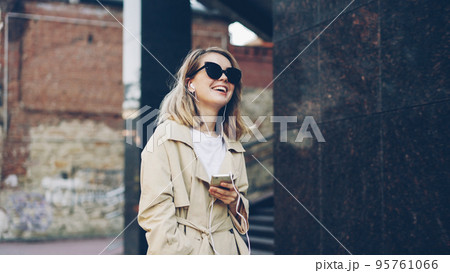 Pretty girl is listening to music through earphones using smartphone and dancing walking in the street and enjoying rhythm and modern city. Girl is wearing sunglasses and trendy coat. 95761066