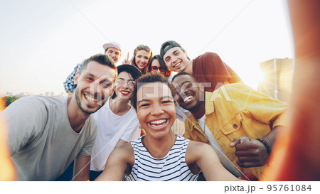 Point of view shot of pretty African American girl holding camera and taking selfie with happy friends at party on roof. Men and women are looking at camera, posing and laughing. 95761084