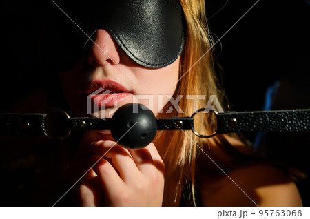 A man dominates and puts a gag in her mouth. BDSM concept. Portrait of a woman in seductive underwear with an intimate toy in her mouth. Sexy couple plays love games. Eyes closed with a leather mask. 95763068