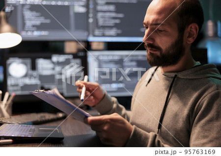Software developer taking notes on files after comparing html script and source code, doing online database analysis. Writing coding information on papers, working late at night in office. Software developer taking notes on files after comparing html script and source code, doing online database analysis. Writing coding information on papers, working late at night in office. 95763169