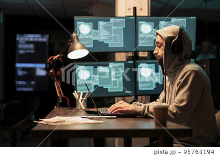 Male criminal trying to break database server, using virus to code malware and steal big data at night. Working with multiple monitors and hacking information system, wearing hood. Male criminal trying to break database server, using virus to code malware and steal big data at night. Working with multiple monitors and hacking information system, wearing hood. 95763194