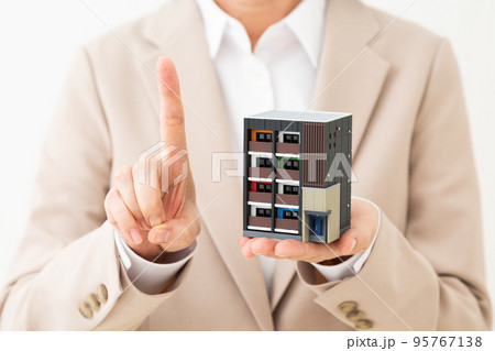 マンションの住宅模型を持って人差し指を立てるスーツ姿のミドル女性 95767138