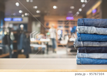 Blue denim jeans stack on wood table top with abstract blur clothing store in modern shopping mall defocused bokeh light background 95767304