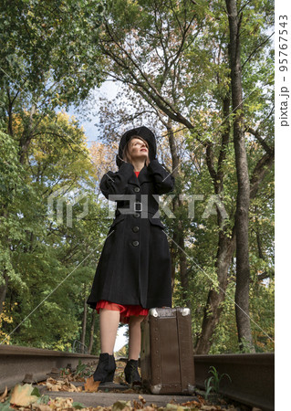 Middle-aged woman in long coat and black hat with brims looks up at sky. Madam stands on rails with suitcase in forest 95767543
