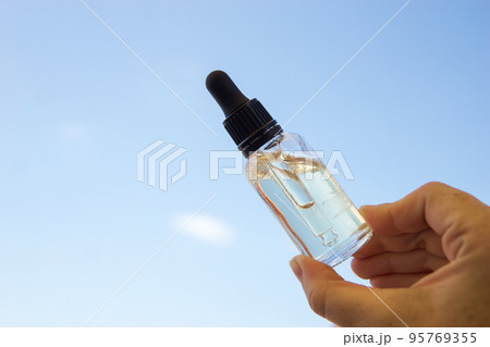 A bottle of cosmetic serum or gel in a woman's hand against a background of blue sky, clouds and sun. The concept of summer holidays and the use of sunscreens 95769355