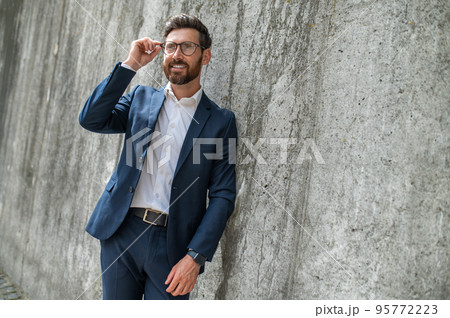 An elegant young man in eyeglasses standing near the wall 95772223