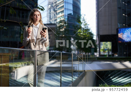 Corporate woman in beige suit, drinks morning coffee, looks at her smartphone screen, uses application, checking messages on phone, stands on street outside office 95772459