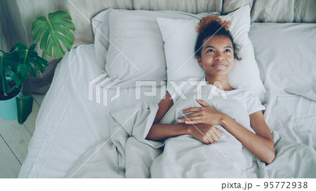 Happy young woman is lying in bed awake and smiling enjoying carefree life, comfortable bed and good news. Positive emotions, bedtime and glad youth concept. 95772938