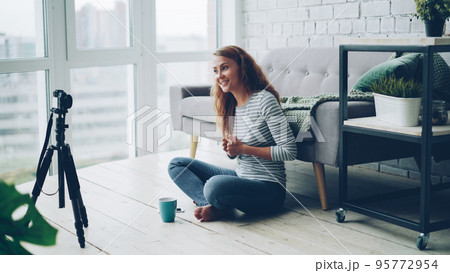 Beautiful young woman vlogger is talking and gesturing recording video with professional camera for internet blog. Girl is wearing casual clothing and sitting on the floor at home. 95772954