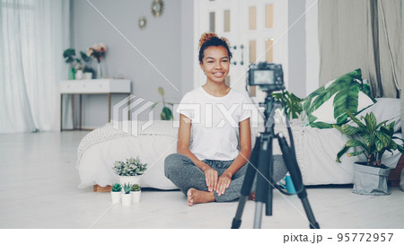 Good-looking African American girl creative blogger is recording video about plants sitting on floor of her modern apartment and talking looking at camera on tripod. 95772957