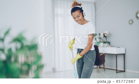 Beautiful African American teenager is mopping floor, dancing and singing listening to radio in wireless headphones. Nice house interior and furniture is visible. Beautiful African American teenager is mopping floor, dancing and singing listening to radio in wireless headphones. Nice house interior and furniture is visible. 95772987