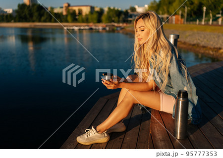 Happy relaxed blonde young woman holding cup with hot coffee from thermos sitting on beach by wooden pier on sunny summer morning. Beautiful female traveler enjoying with nature drinking tea over lake 95777353