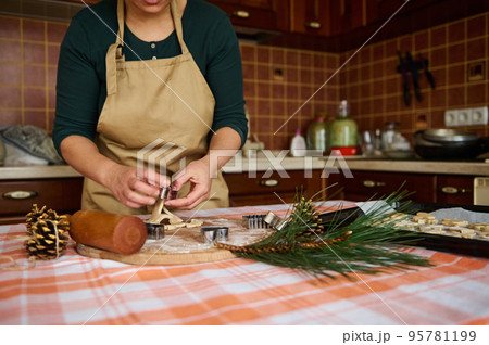 A woman housewife, wearing a green shirt and a beige chef's apron, using cookie cutters, preparing gingerbread cookies of various shapes, for Christmas in the kitchen at home. Pine cones as ornament 95781199