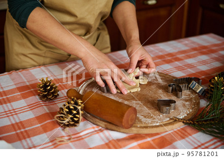 Close-up of a woman housewife, wearing a beige chef's apron, preparing homemade gingerbread cookies for Christmas in the kitchen at home. Female confectioner using cookie cutter, making biscuits 95781201