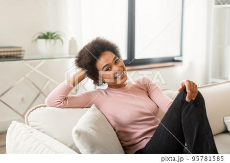 Cheerful relaxed young african american woman with curly hair dream, sits on sofa enjoys silence Cheerful relaxed young african american woman with curly hair dream, sits on sofa enjoys silence 95781585