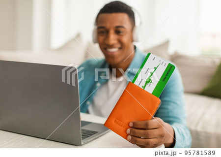 Happy young african american man in wireless headphones shows passport and air tickets, looks at computer Happy young african american man in wireless headphones shows passport and air tickets, looks at computer 95781589