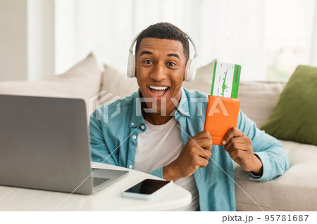 Glad excited young african american guy in wireless headphones with passport, air tickets, near computer Glad excited young african american guy in wireless headphones with passport, air tickets, near computer 95781687
