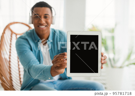 Happy young african american man with wireless headphones showing tablet with blank screen Happy young african american man with wireless headphones showing tablet with blank screen 95781718