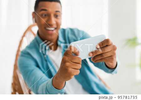 Glad excited young african american man with wireless headphones plays online game on smartphone 95781735
