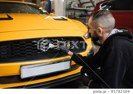 Male worker in gloves washes car radiator grille of luxury yellow car with special brush and soap 95782309