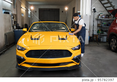 Wash worker man, wearing protective clothes uniform overalls , cleaning modern yellow luxury car 95782397