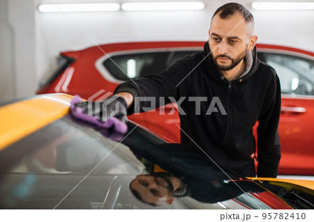 Car wash worker man, wearing protective gloves, cleaning modern car window with microfiber Car wash worker man, wearing protective gloves, cleaning modern car window with microfiber 95782410