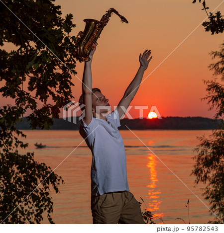 A young man with a saxophone raised his hands up, enjoying the sunset. A young man with a saxophone raised his hands up, enjoying the sunset. 95782543
