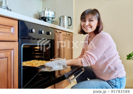 Middle-aged woman with cooked meat taking baking sheet out of oven Middle-aged woman with cooked meat taking baking sheet out of oven 95783589