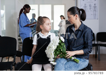 Mom, daughter waiting to see hospitalized family member in hospital reception. Girl, young woman in hospital visiting area. Relatives bringing flowers to hospital patient. 95783789