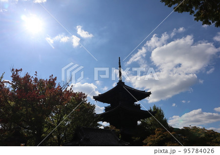 白く輝く太陽と青空と白い雲と色づく樹と緑の木と三重塔のあるお寺の境内の風景 95784026