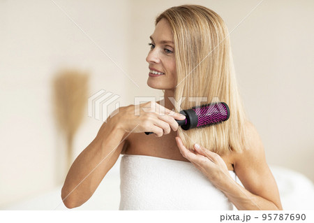 Beautiful middle aged woman in bath towel combing her smooth hair with modern brush, standing in bathroom interior 95787690