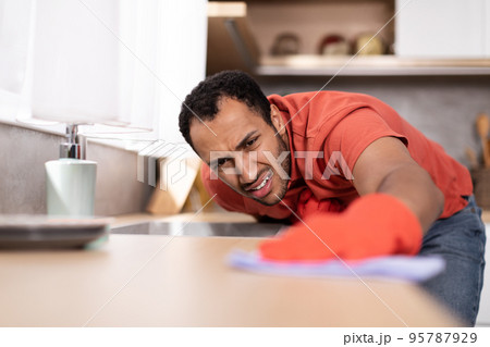 Sad dissatisfied young black male in red t-shirt wipes dirt from wooden table in kitchen interior, close up 95787929