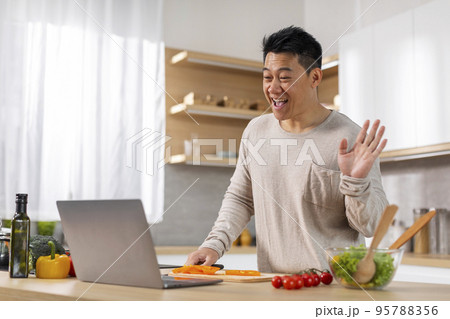 Asian man cooking dinner, having video conversation, using laptop Asian man cooking dinner, having video conversation, using laptop 95788356
