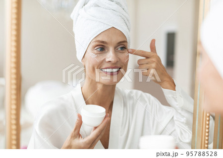 Beautiful middle aged woman in silky bathrobe applying moisturizing eye cream, enjoying face care routine Beautiful middle aged woman in silky bathrobe applying moisturizing eye cream, enjoying face care routine 95788528