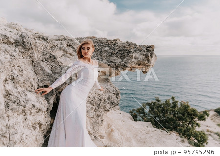 Romantic bride, blond girl in white wedding dress with open shoulders posing with sea and rocks in background. Stylish young woman standing on cliff's edge and looking into a ocean mountains at sunset 95795296