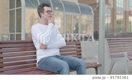 Pensive Young Man Thinking while Sitting Outdoor on Bench Pensive Young Man Thinking while Sitting Outdoor on Bench 95795361