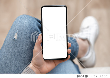 Top view mockup image of woman holding mobile phone with blank white screen. Woman in jeans sits cross-legged and holds cell phone in her hands with blank white screen Top view mockup image of woman holding mobile phone with blank white screen. Woman in jeans sits cross-legged and holds cell phone in her hands with blank white screen 95797382