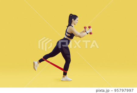 Sportswoman working out with elastics ribbon and with dumbbells on vivid yellow background. 95797997