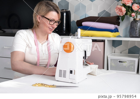 Real middle aged woman tailor sewing clothes using a sewing machine at home. Seamstress working. Hand making clothes in home interior. Female sits enjoying process of sewing clothes. Real middle aged woman tailor sewing clothes using a sewing machine at home. Seamstress working. Hand making clothes in home interior. Female sits enjoying process of sewing clothes. 95798954